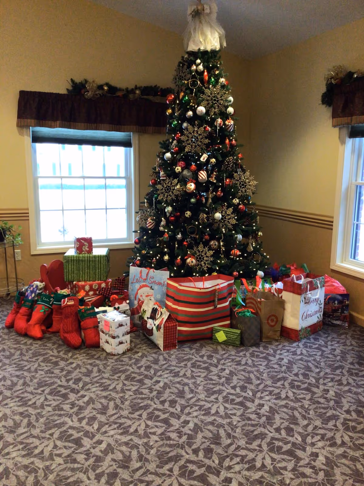A decorated Christmas tree with an angel on top, surrounded by numerous wrapped gifts and Christmas stockings, placed in a room with two windows and patterned carpet.