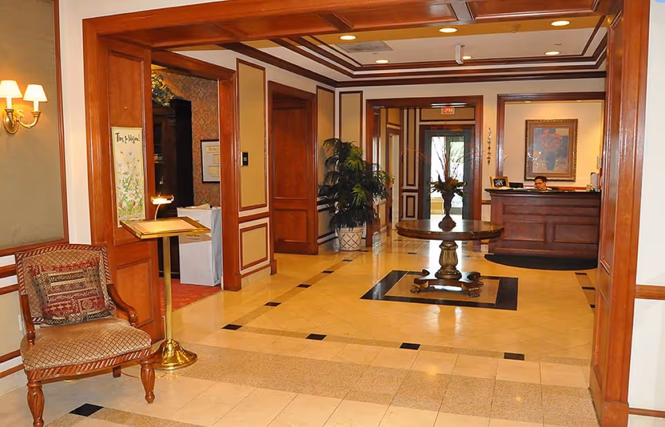 A spacious and elegant lobby area with polished tile flooring and wooden trim. There is a round wooden table with a flower arrangement in the center of the floor. To the right, a reception desk with a person seated behind it is visible. On the left, a cushioned wooden chair with a decorative pillow and a stand with a sign are present. The walls are adorned with framed artwork and sconces with lampshades, and there are potted plants near the back hallway.