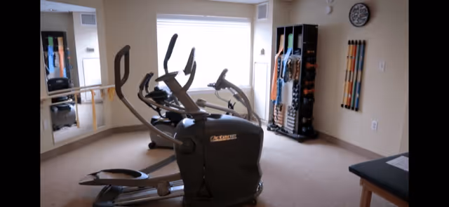 A small exercise room with two elliptical machines positioned near a large window. The room has beige walls and carpeted floor. On the right side, there is a rack holding various exercise equipment such as resistance bands and weights. A clock is mounted on the wall above the rack, and a mirror is visible on the left wall.
