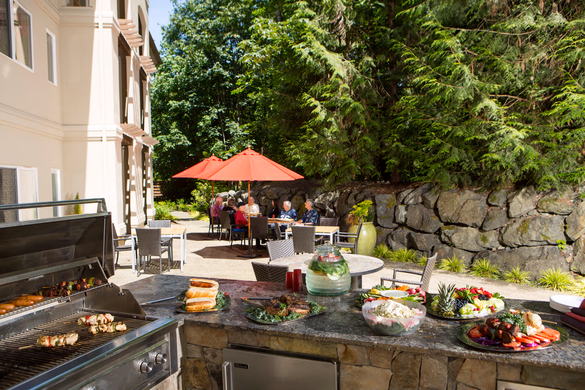 Outdoor patio area at Aegis Living Marymoor with a built-in grill and countertop displaying various foods including skewers, bread, salads, and fruit platters. In the background, several elderly people are seated at tables under red umbrellas, surrounded by greenery and a stone wall.
