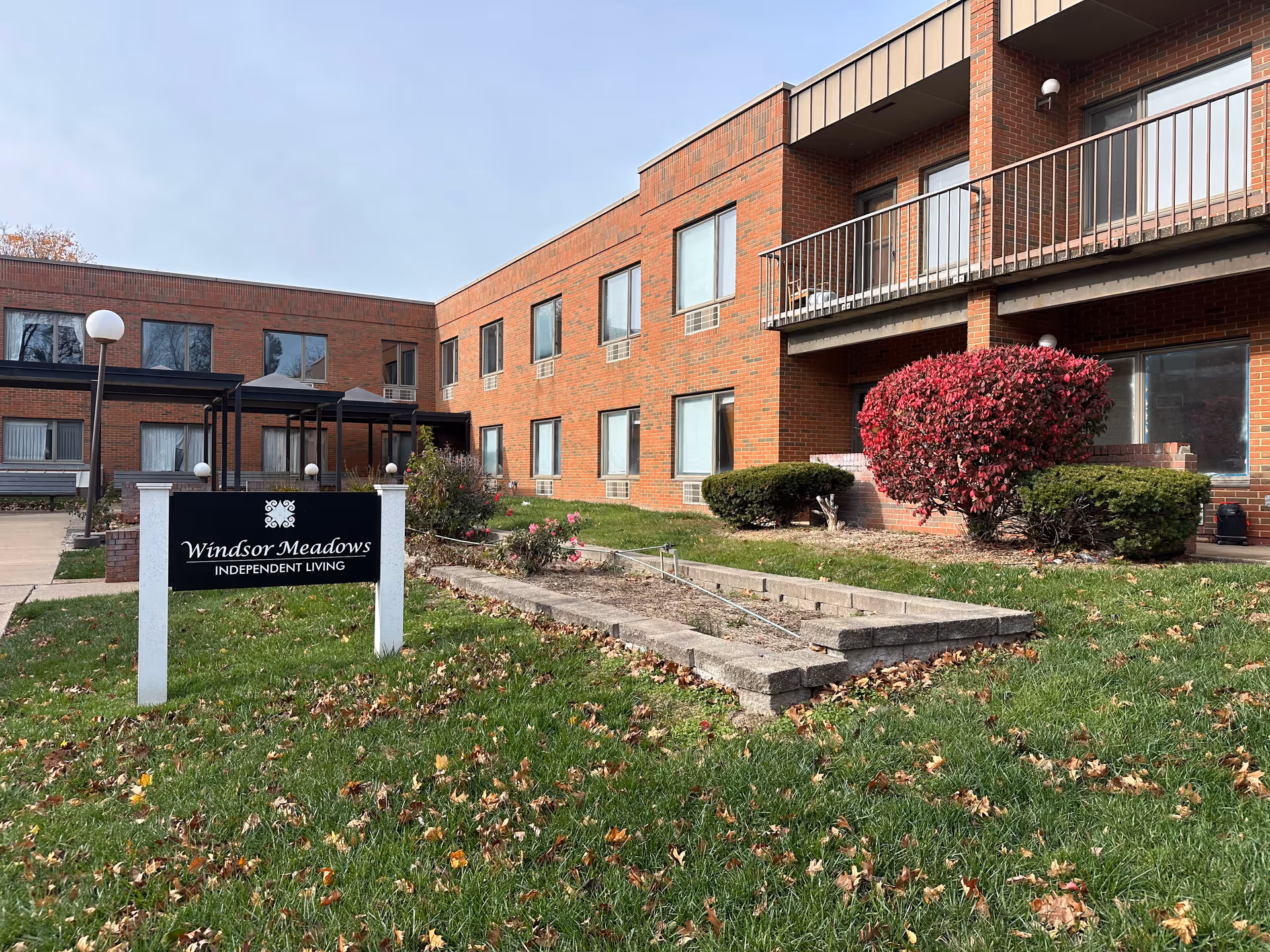 Exterior view of a two-story brick building with multiple windows and balconies. In front of the building is a green lawn with scattered fallen leaves, a small garden area with bushes and flowers, and a black and white sign that reads 'Windsor Meadows Independent Living'. The sky is clear with some light clouds.