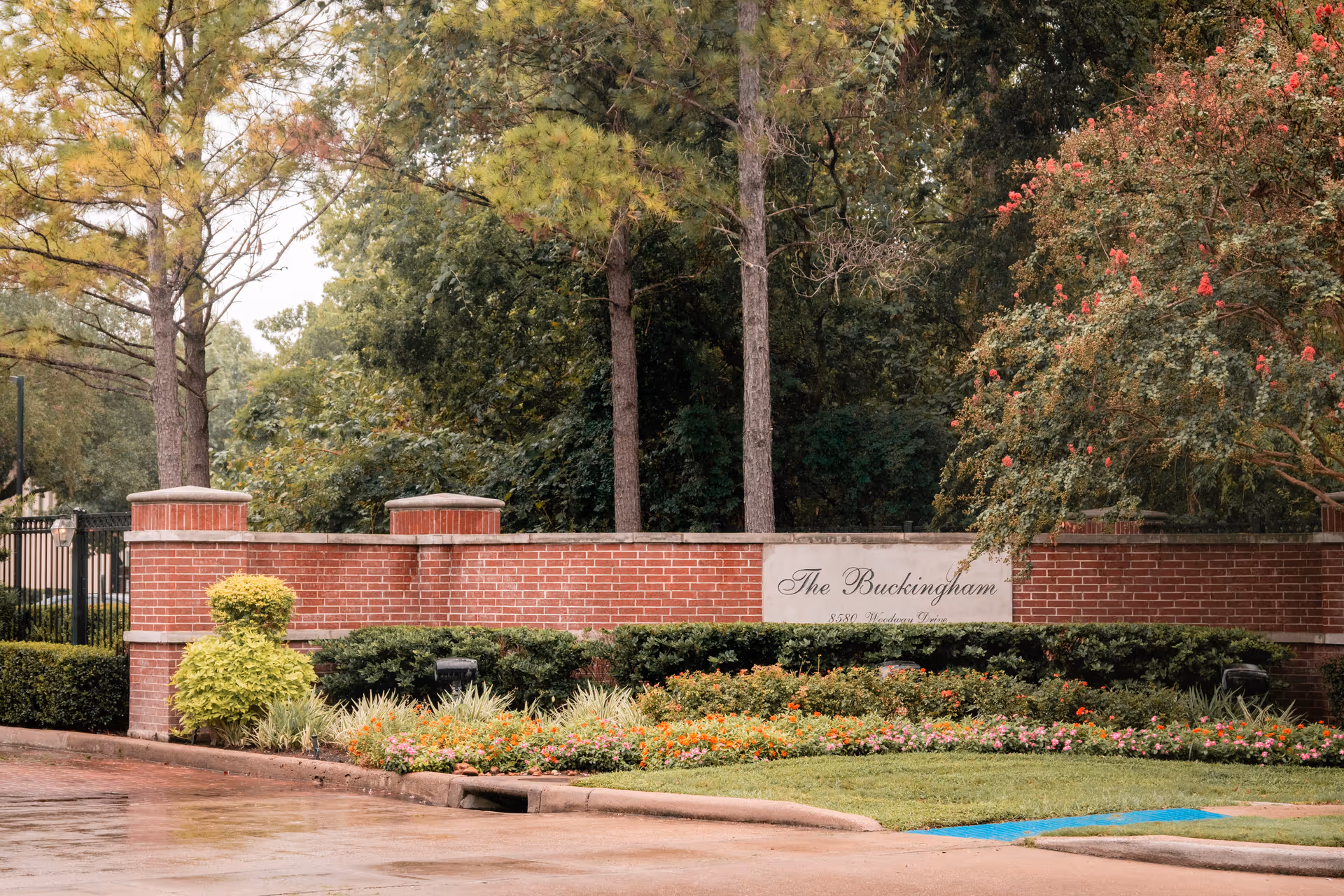 Entrance sign for The Buckingham facility made of red brick with a white plaque displaying the name and address, surrounded by green bushes, colorful flowers, and tall trees in the background.