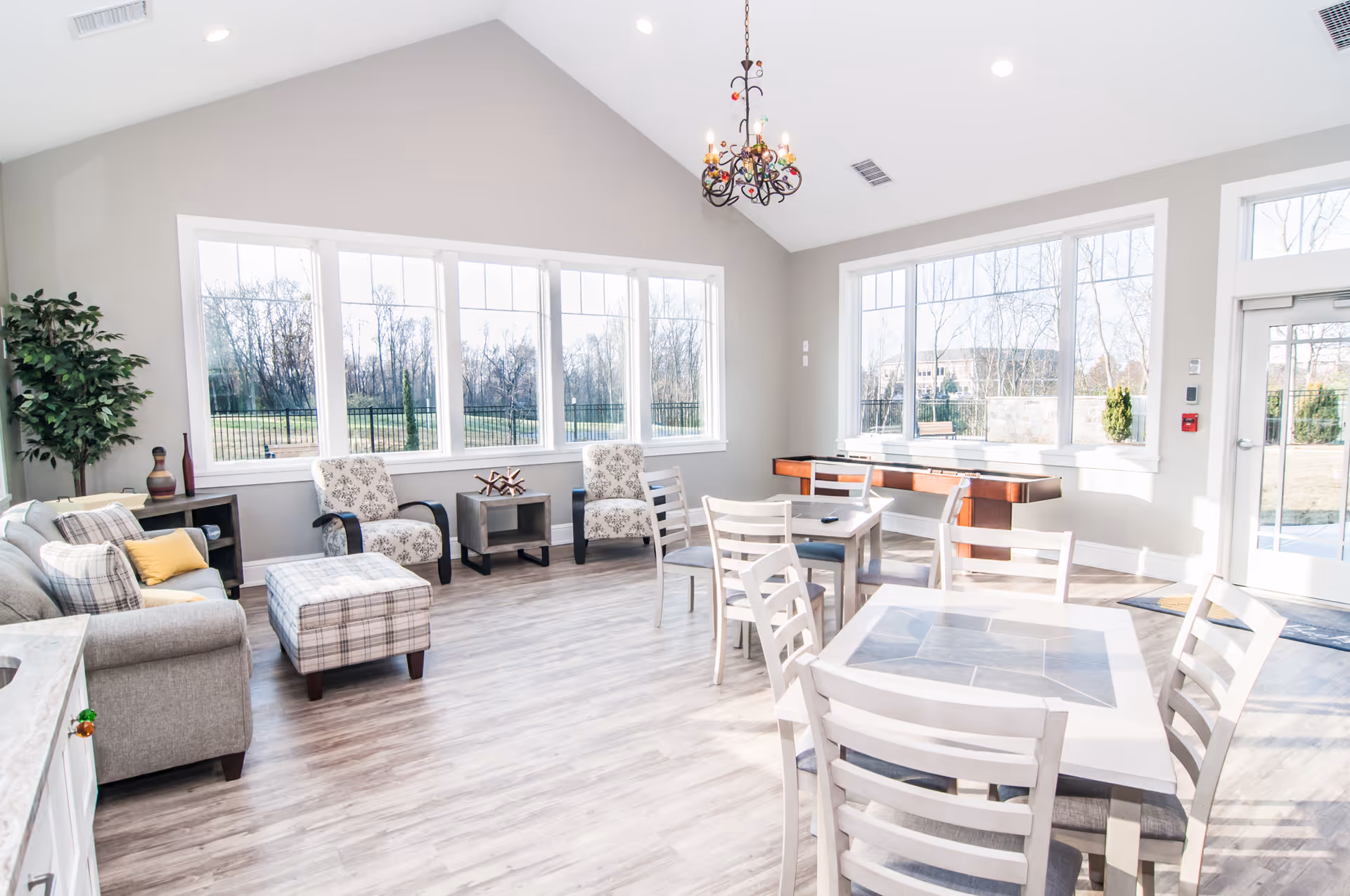 A bright and spacious common area in a senior living facility with large windows letting in natural light. The room features a gray sofa with plaid and yellow pillows, two patterned armchairs, a plaid ottoman, several white wooden tables with matching chairs, a shuffleboard table, and a decorative chandelier hanging from the vaulted ceiling. Outside the windows, trees and a fenced yard are visible.