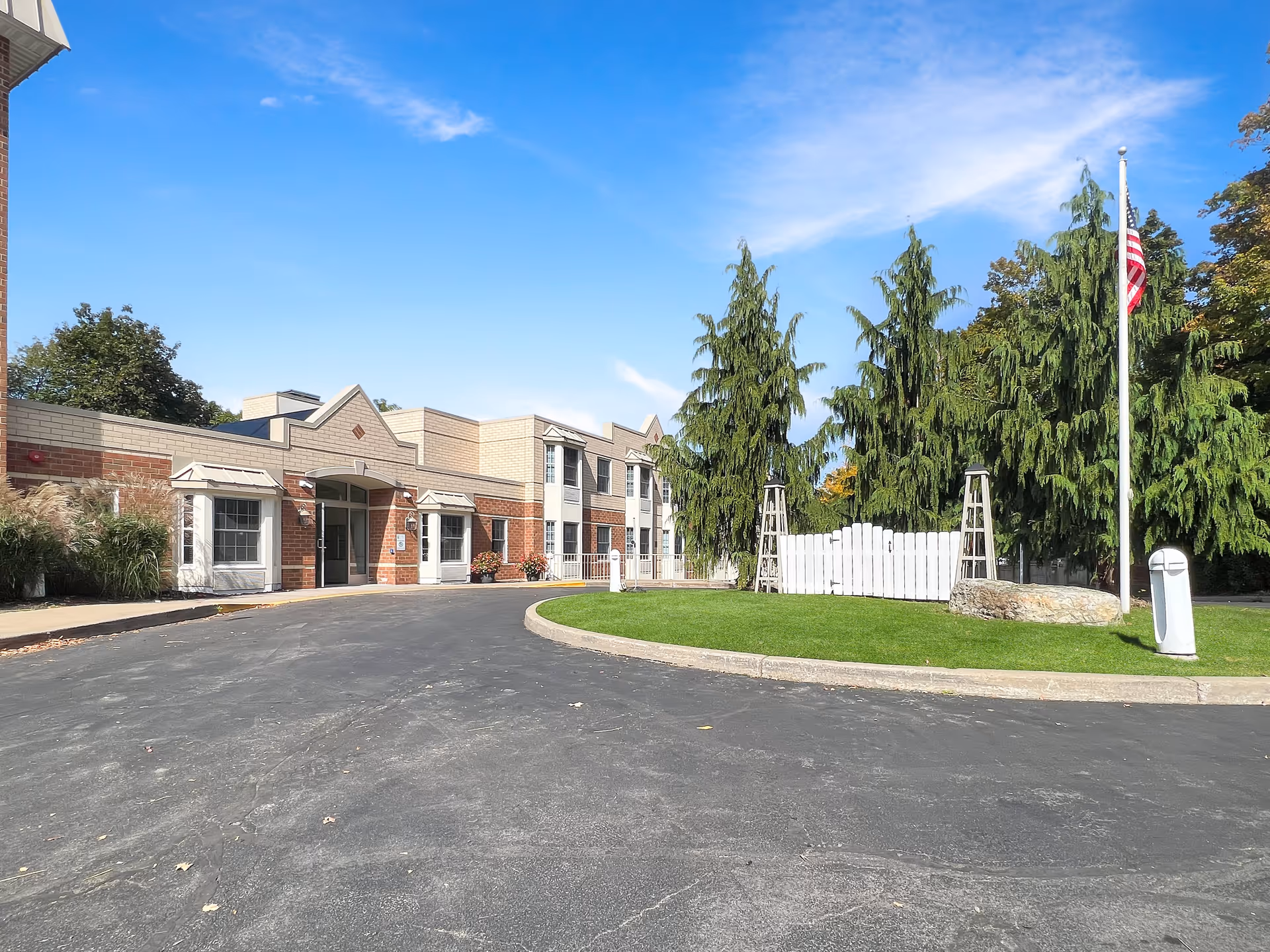 Exterior view of a senior living facility building with a curved driveway, green lawn, tall evergreen trees, and an American flag on a flagpole under a clear blue sky.
