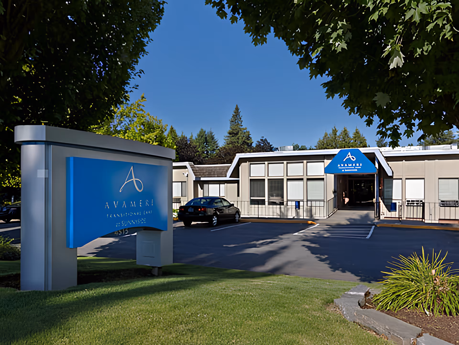 Exterior view of Avamere Transitional Care at Sunnyside building with a blue sign in the foreground and a car parked near the entrance. The building is surrounded by trees and greenery under a clear blue sky.
