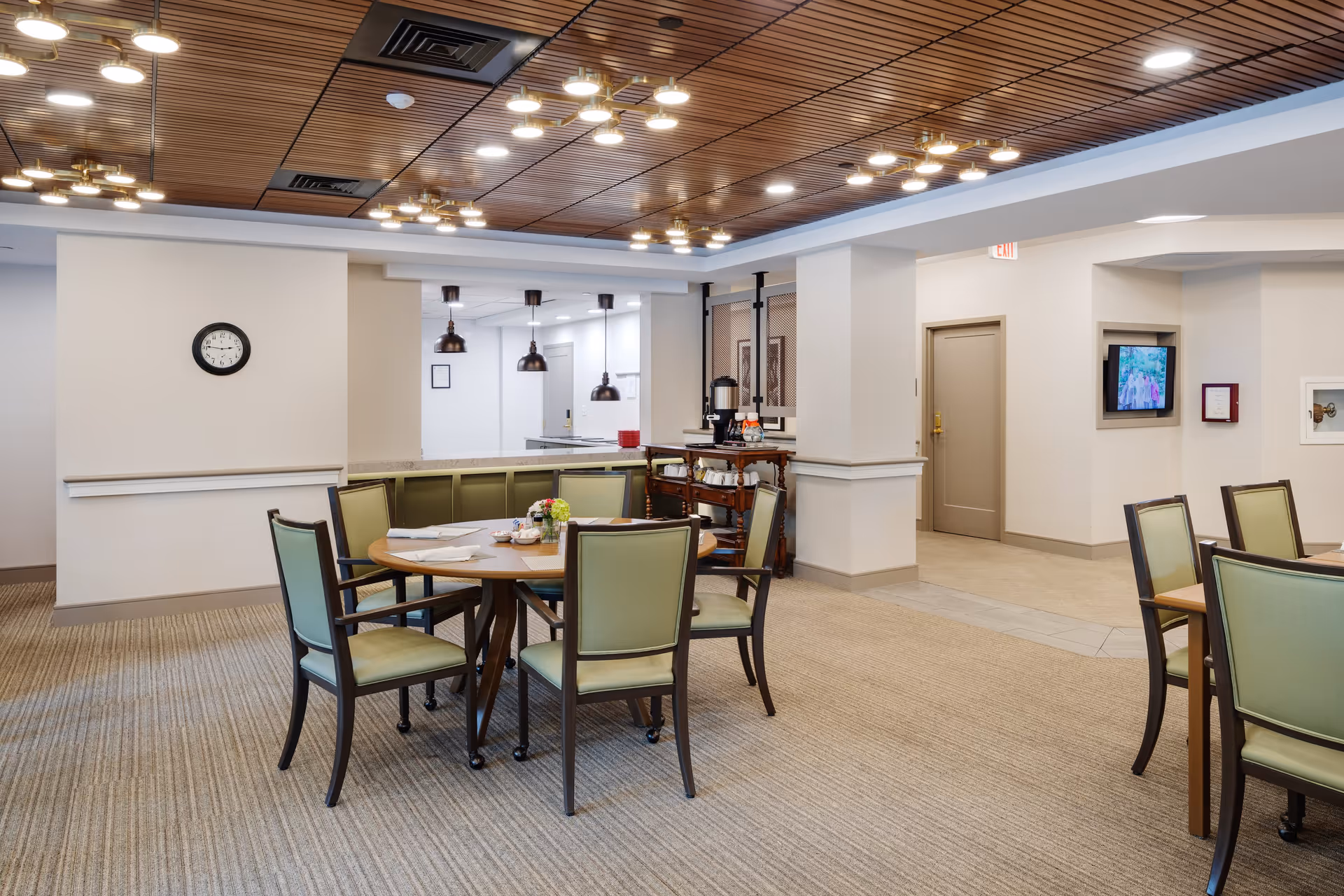 A well-lit dining area in a senior living facility with round wooden tables and green cushioned chairs. The ceiling has wooden paneling with multiple modern light fixtures. There is a clock on the wall, a small service cart with coffee supplies, and a TV mounted on the wall in the background.