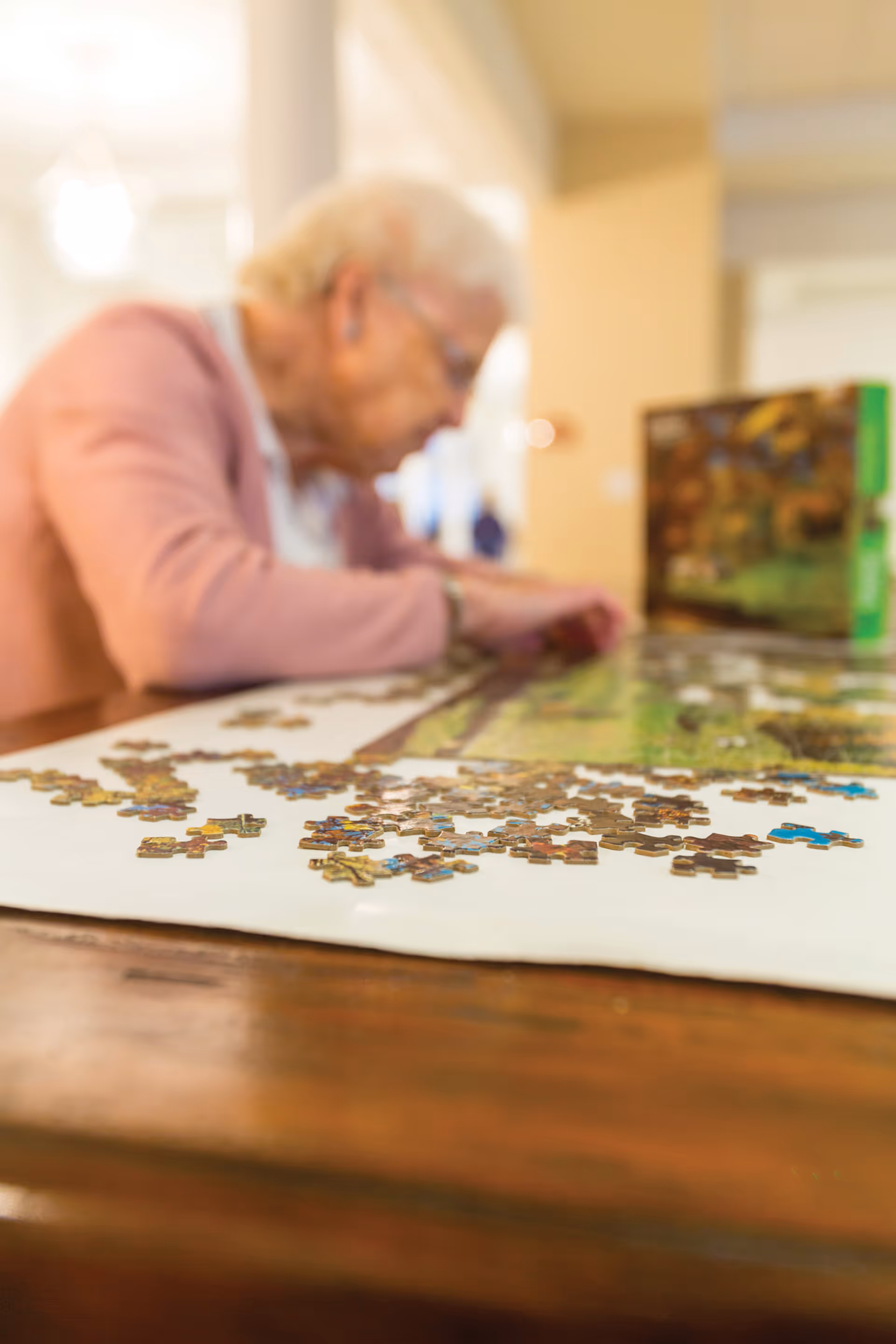 An elderly woman wearing a pink cardigan is working on assembling a jigsaw puzzle on a wooden table indoors. The puzzle pieces are spread out on a white surface, and the woman is focused on placing pieces together.