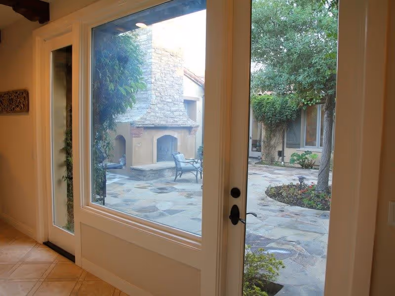 View through glass doors from inside a building looking out onto a stone patio with an outdoor fireplace, chairs, trees, and plants.