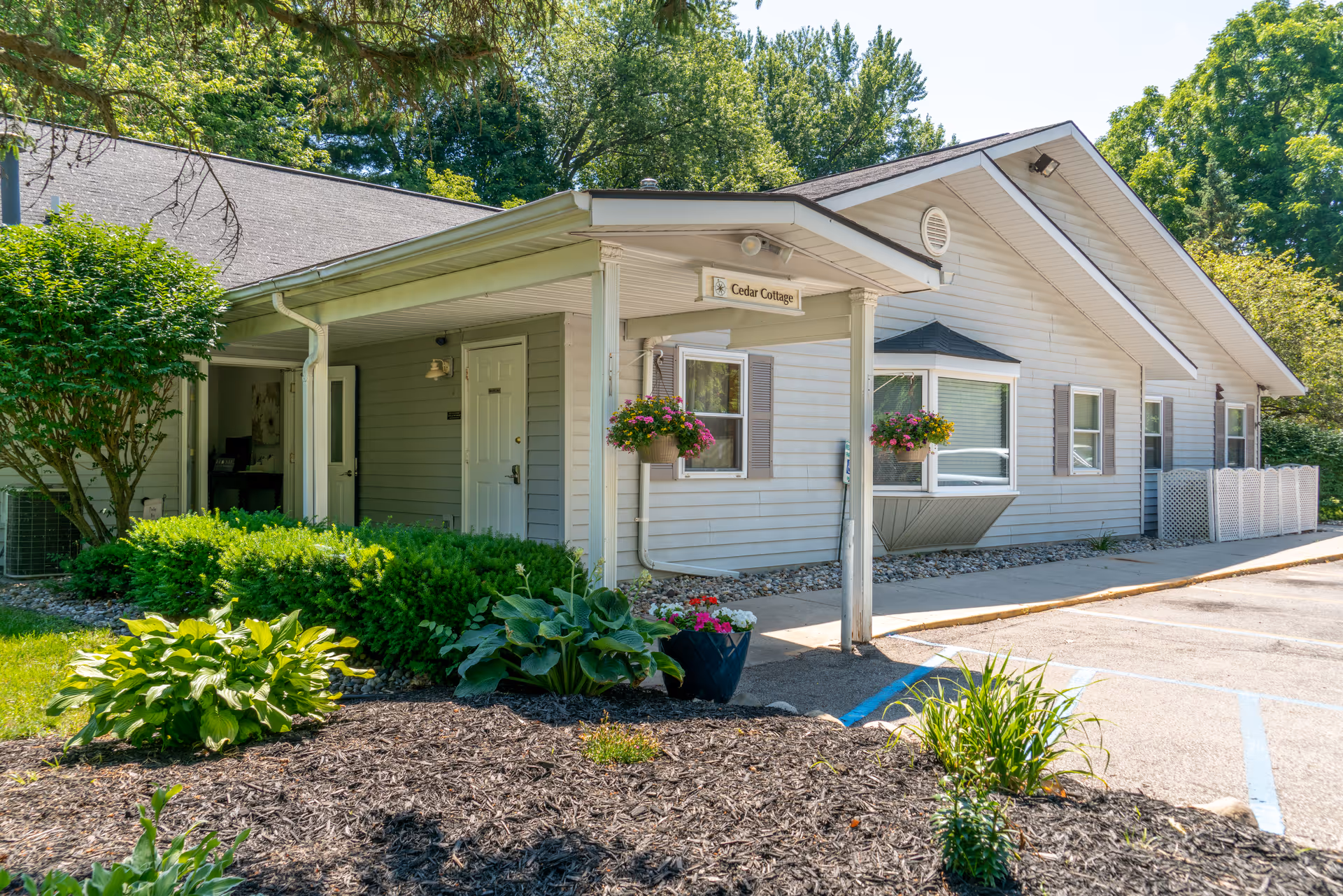 Exterior view of a single-story building named Cedar Cottage with light gray siding, white trim, and a covered entrance. The building is surrounded by green shrubs, plants, and trees, with a paved parking area visible on the right side. Hanging flower pots decorate the entrance area.