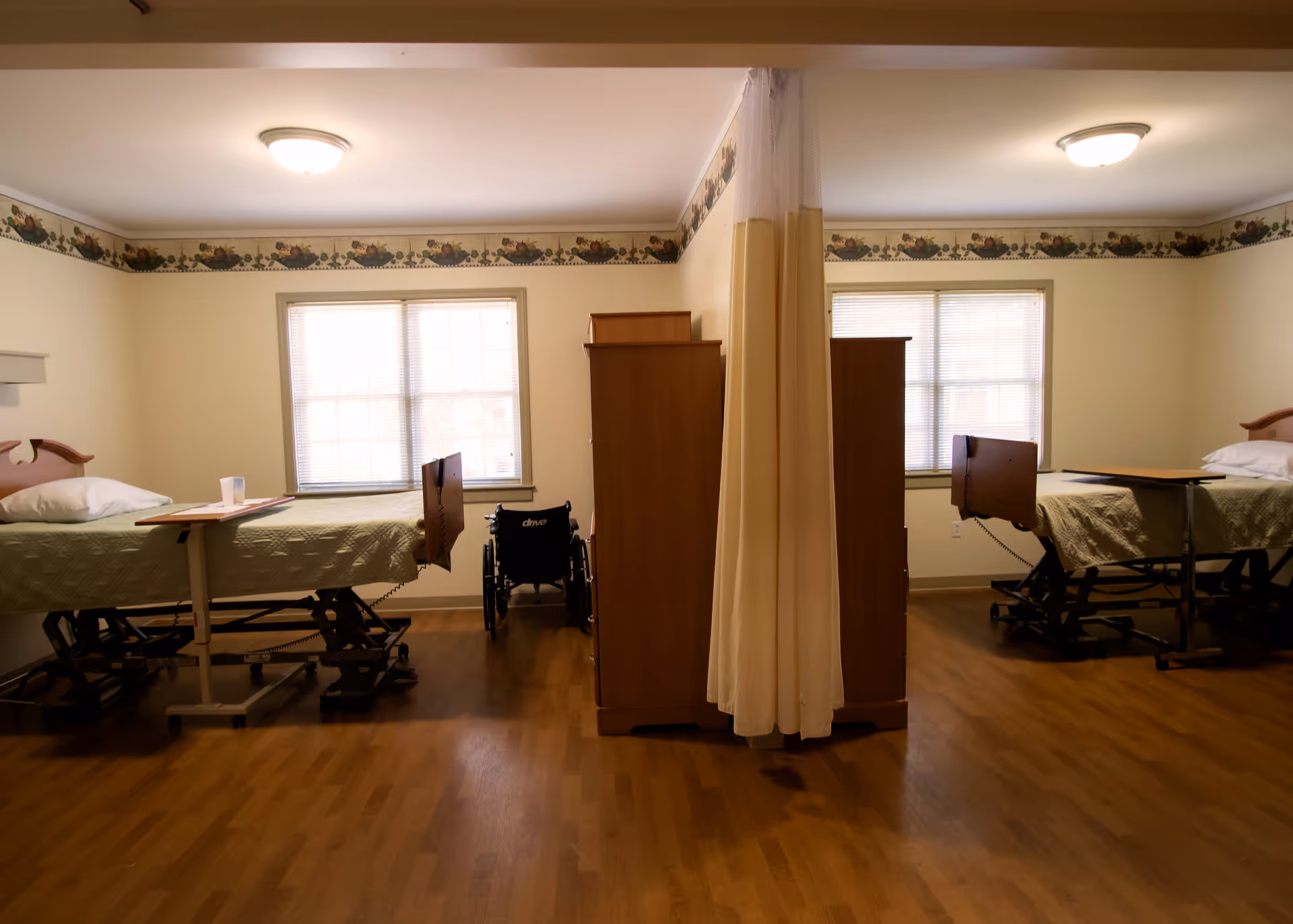 A shared nursing room with two hospital beds separated by a privacy curtain. Each bed has a green quilted blanket, a pillow, and an overbed table. There is a wheelchair positioned near the window between the beds. The room has wooden flooring, two windows with blinds, and a floral border near the ceiling.