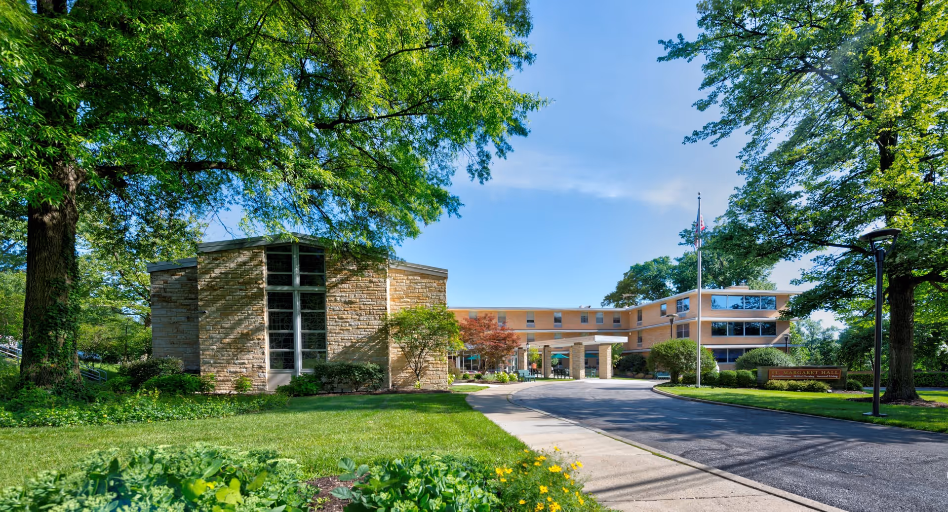 Exterior view of CareCore at Margaret Hall facility showing a multi-story building with large windows, a covered entrance, a flagpole with an American flag, and well-maintained green landscaping with trees and shrubs under a clear blue sky.