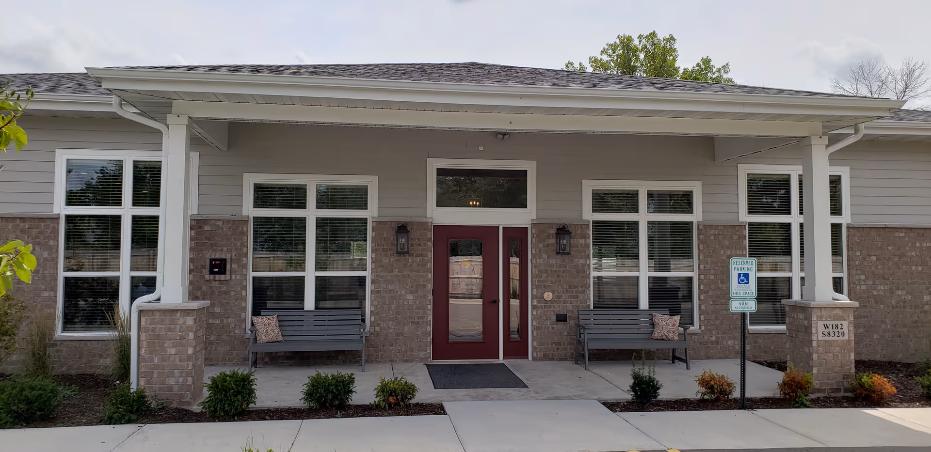 Front exterior view of a single-story building with a covered entrance featuring double red doors, two benches with pillows on either side, large windows with white blinds, brick and siding facade, and a reserved parking sign for handicapped accessible parking.
