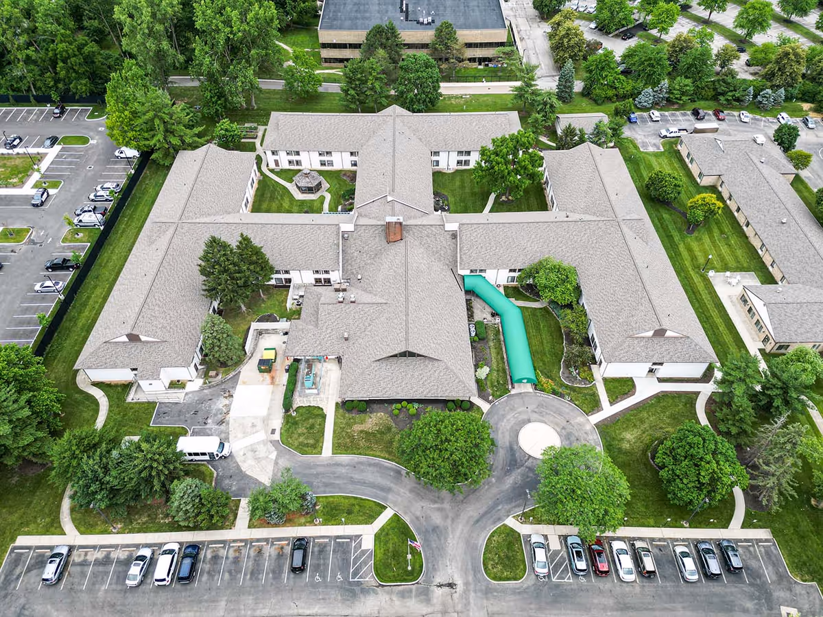 Aerial view of The Village of Westerville senior living facility showing multiple connected buildings with gray roofs, surrounded by green lawns, trees, and parking lots with several cars parked.