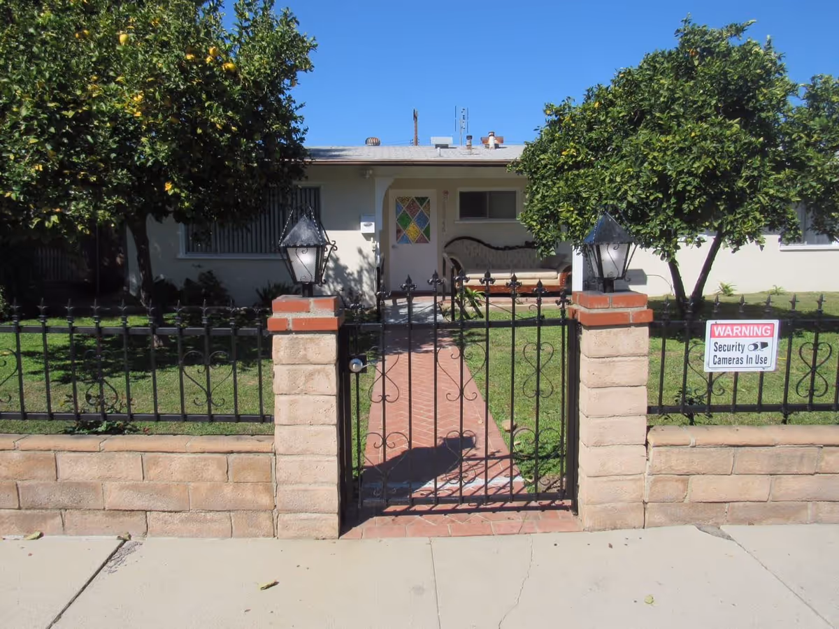Front view of a single-story house with a small front yard enclosed by a black wrought iron fence and gate. There are two trees with green foliage on either side of a brick pathway leading to the front door, which has a colorful stained glass window. Two lantern-style lights are mounted on brick pillars flanking the gate. A sign on the fence reads 'WARNING Security Cameras In Use.'