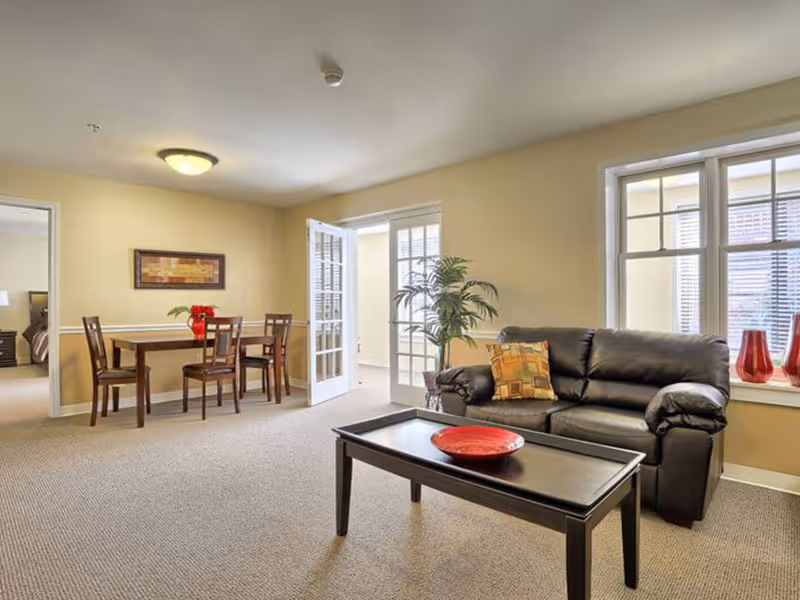 A bright living room area with beige walls and carpeted floor. The room features a black leather sofa with a patterned cushion, a dark wooden coffee table with a red decorative bowl, and a dining table with four chairs. There is a potted plant near the window, and French doors leading to another room. The adjacent room has a bed visible through an open doorway.