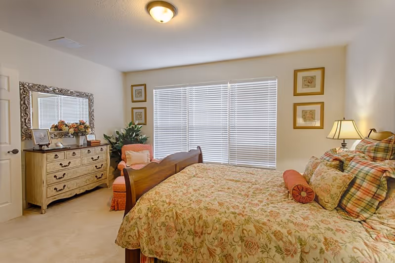 A cozy bedroom with a wooden bed frame and floral patterned bedding. There is a wooden dresser with a large ornate mirror above it, decorated with a flower arrangement and framed photos. A coral-colored armchair with a cushion is placed next to the dresser. The room has beige walls, carpeted floor, and a window with white blinds letting in natural light. Two framed pictures hang on the wall beside the window, and a table lamp is on a nightstand next to the bed.