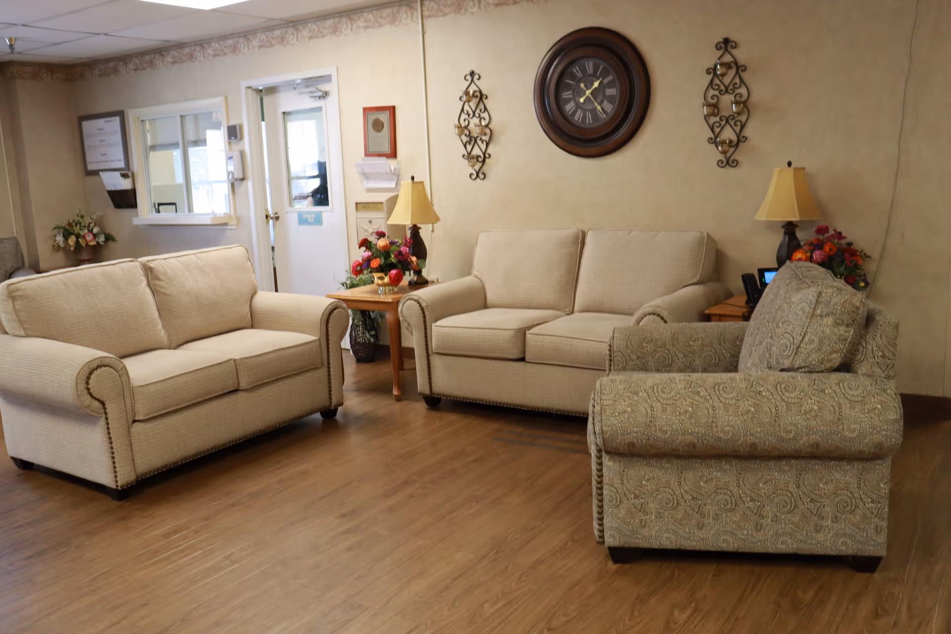 A cozy seating area in a nursing facility with two beige sofas and one patterned armchair arranged around two wooden side tables with lamps and flower arrangements. A large round wall clock and decorative wall hangings are on the beige wall behind the seating.