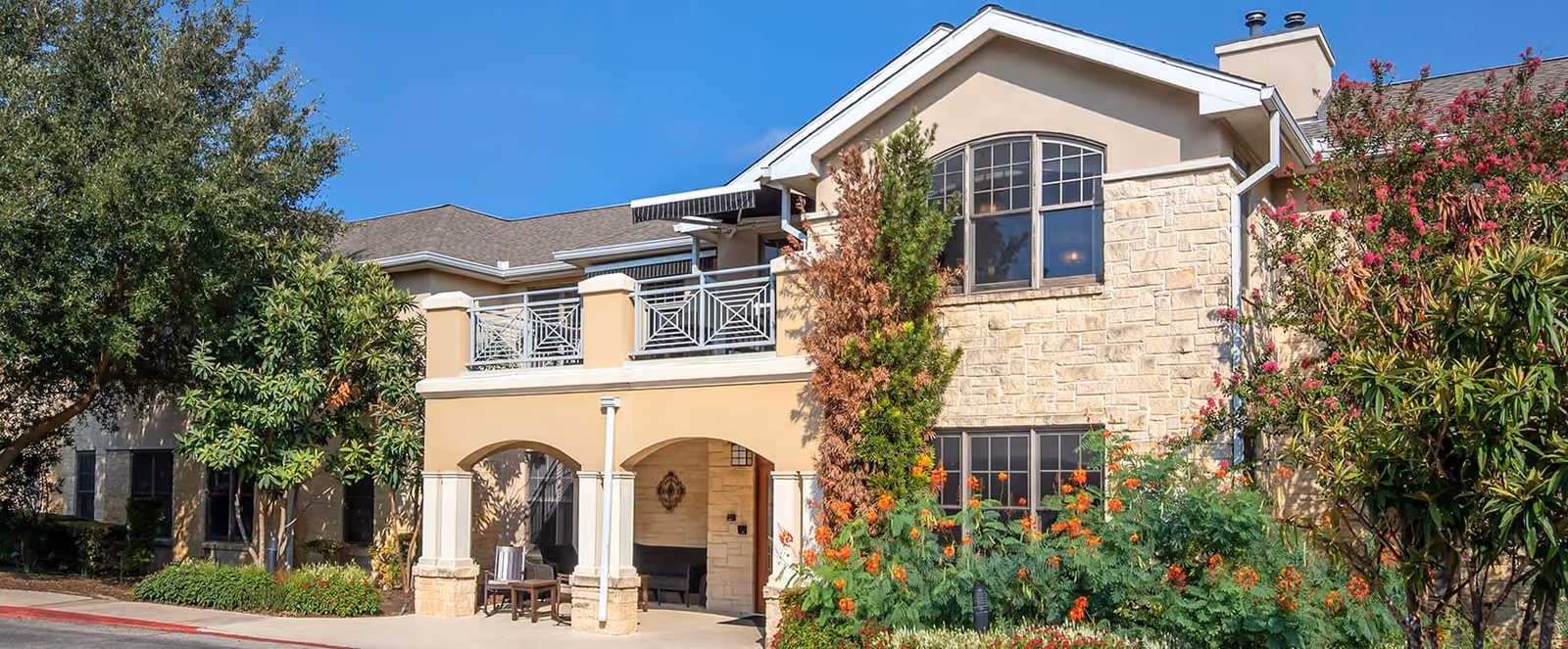 Exterior view of a senior living facility building with stone and beige stucco walls, large windows, a covered entrance with columns, outdoor seating, and lush greenery including trees and flowering plants under a clear blue sky.