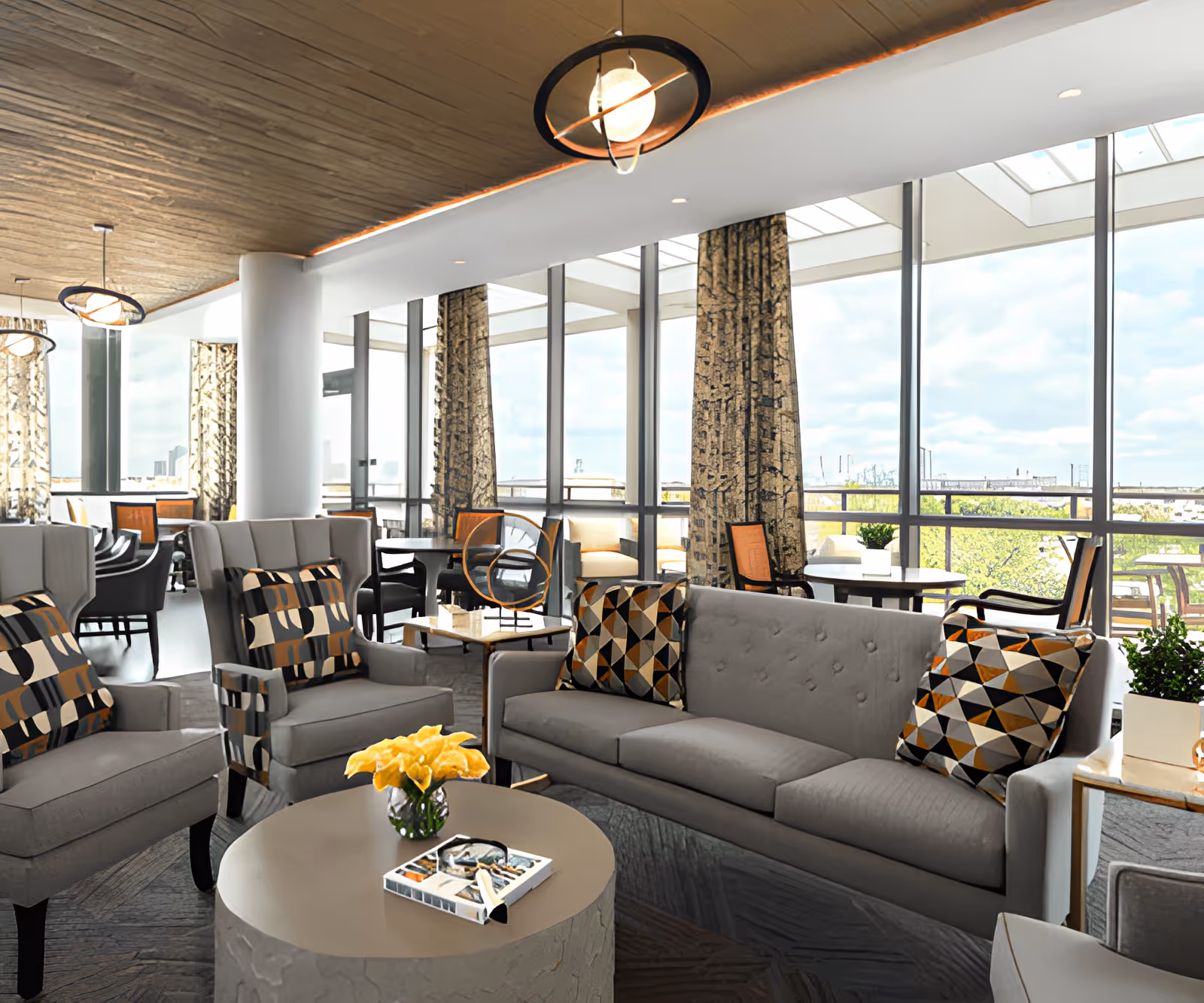 A modern senior living facility lounge area with gray upholstered sofas and armchairs featuring geometric patterned cushions. A round coffee table with a small vase of yellow flowers and a book is in the center. Large floor-to-ceiling windows with patterned curtains allow natural light and offer a view of the outdoors. Additional seating and tables are visible in the background, along with contemporary ceiling lights.