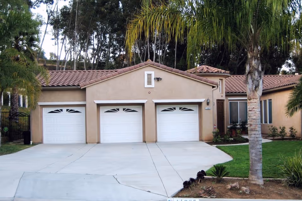 Front exterior view of a single-story residential building with three white garage doors, beige stucco walls, a tiled roof, and surrounding palm trees and greenery.
