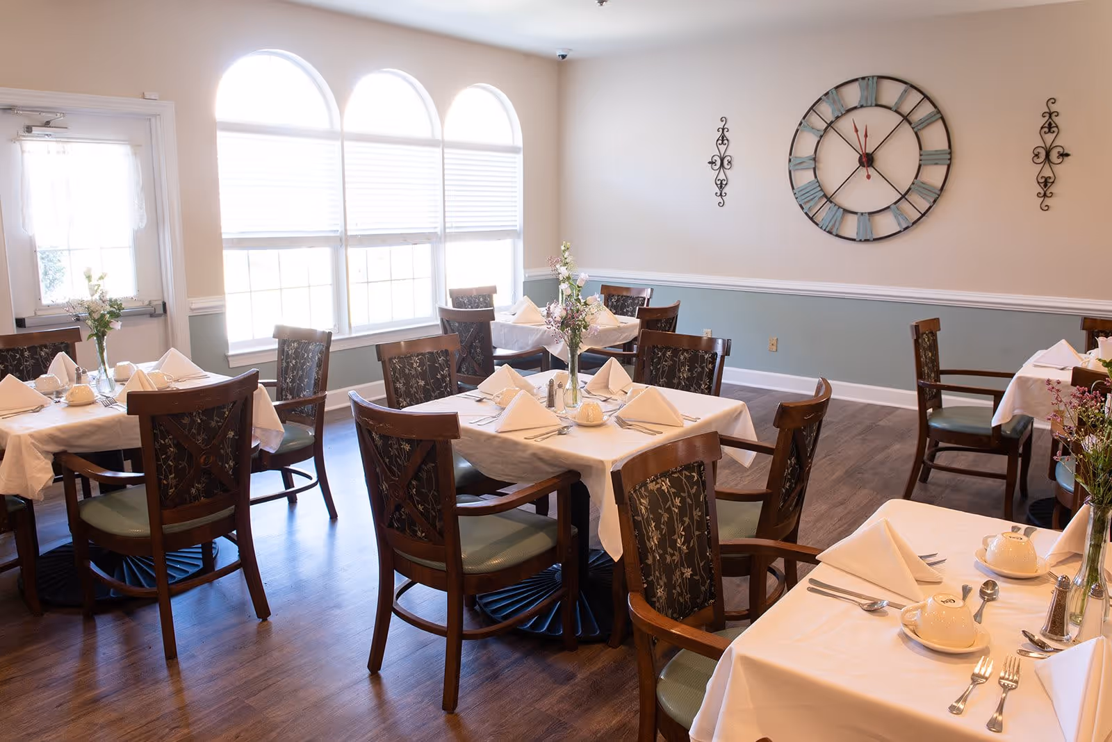 A bright dining room with several tables covered in white tablecloths, each set with folded napkins, cups, and silverware. Wooden chairs with patterned backs surround the tables. Large arched windows allow natural light to fill the room. A large decorative wall clock and ornamental wall hangings are visible on the beige and light blue walls.