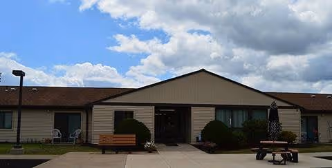 Single-story building with beige siding and a brown roof under a partly cloudy sky. There is a concrete walkway leading to the entrance, flanked by bushes and outdoor seating including a bench and a picnic table with a closed umbrella.