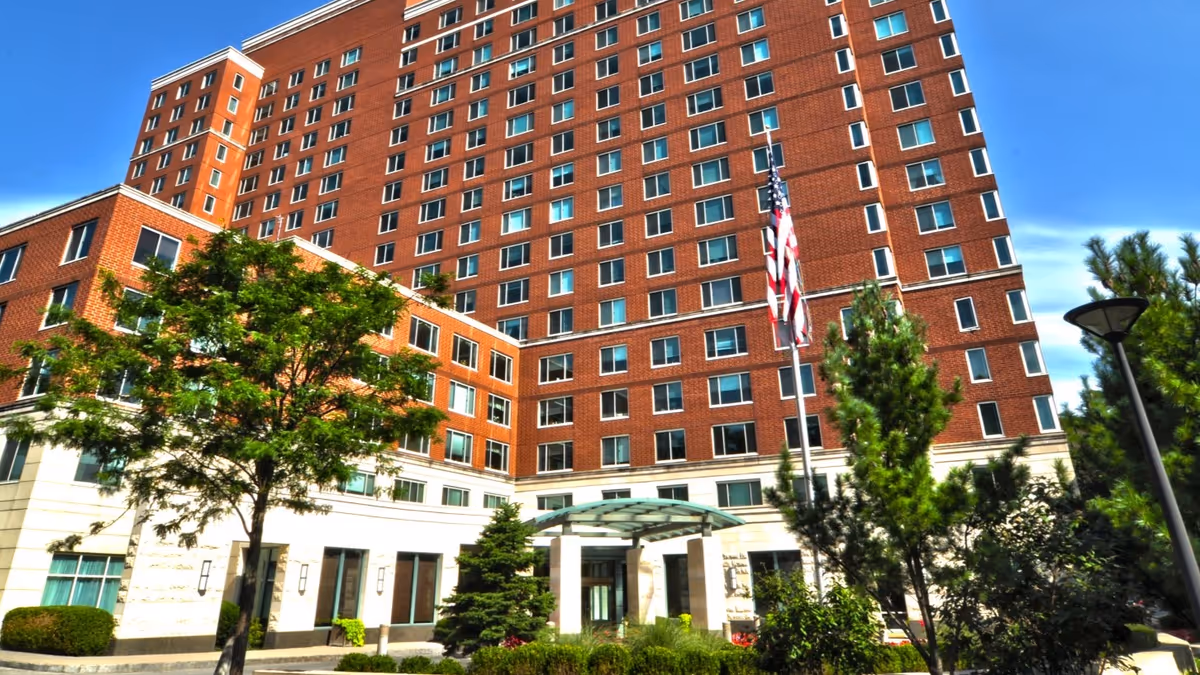 Exterior view of a tall multi-story brick building with many windows, surrounded by trees and greenery under a clear blue sky. The entrance features a glass canopy and an American flag is displayed on a flagpole near the entrance.