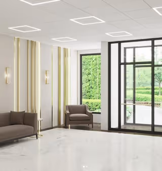 A modern, spacious lobby area with white marble flooring, beige walls with vertical gold accents, a brown sofa and armchair, and large glass doors and windows showing green outdoor landscaping.