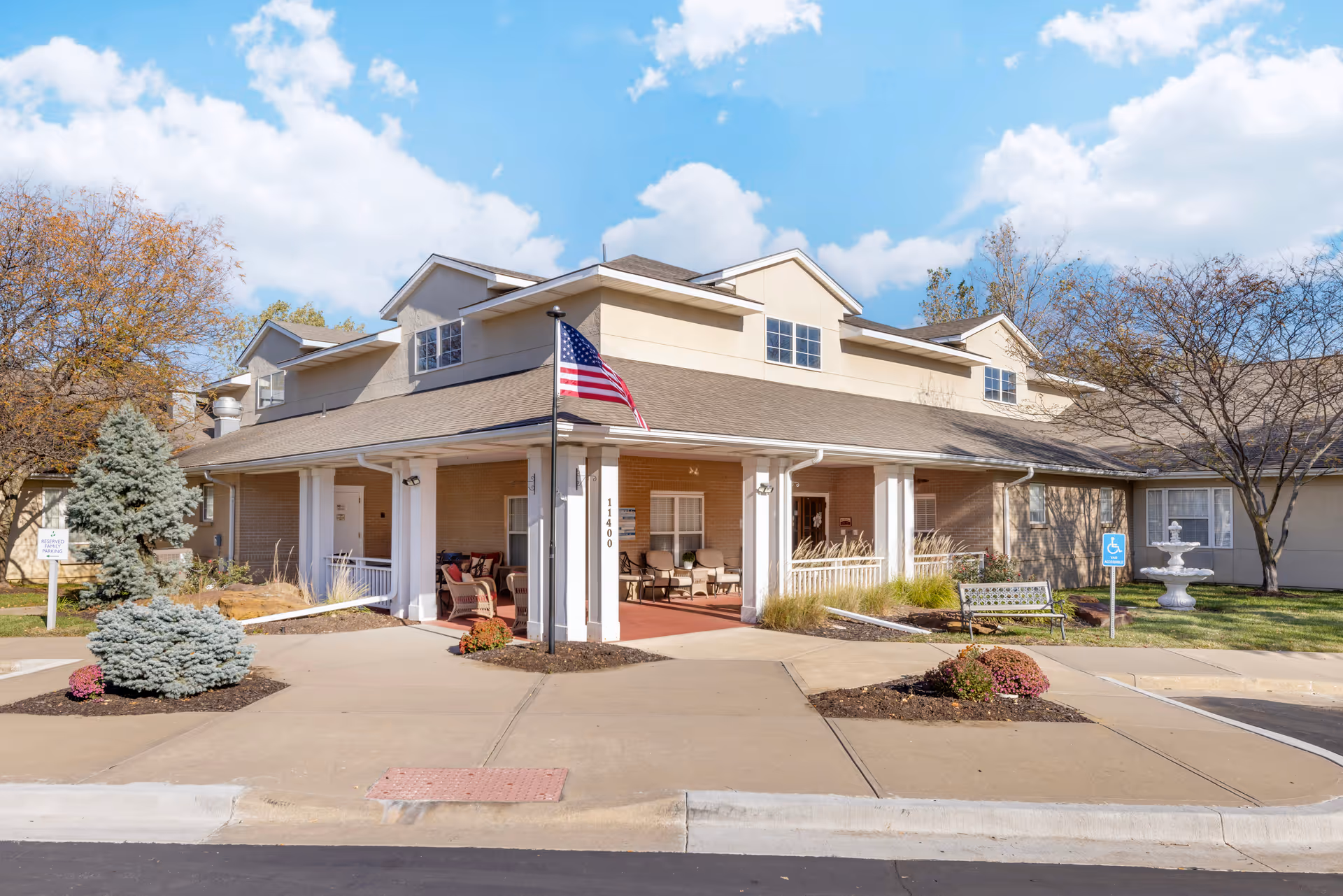 Exterior front view of Brookdale Shawnee facility showing a beige building with a covered entrance supported by white columns. There is an American flag on a pole in front, outdoor seating under the covered area, landscaped bushes, a bench, a white fountain, and a blue sky with clouds.