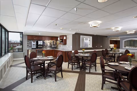 A bright dining room with several round tables set with plates and glasses, surrounded by wooden chairs with cushioned seats. The room features large windows on one side allowing natural light, a beverage station with coffee machines and cabinets, and neutral-colored walls and carpet with a modern ceiling design.