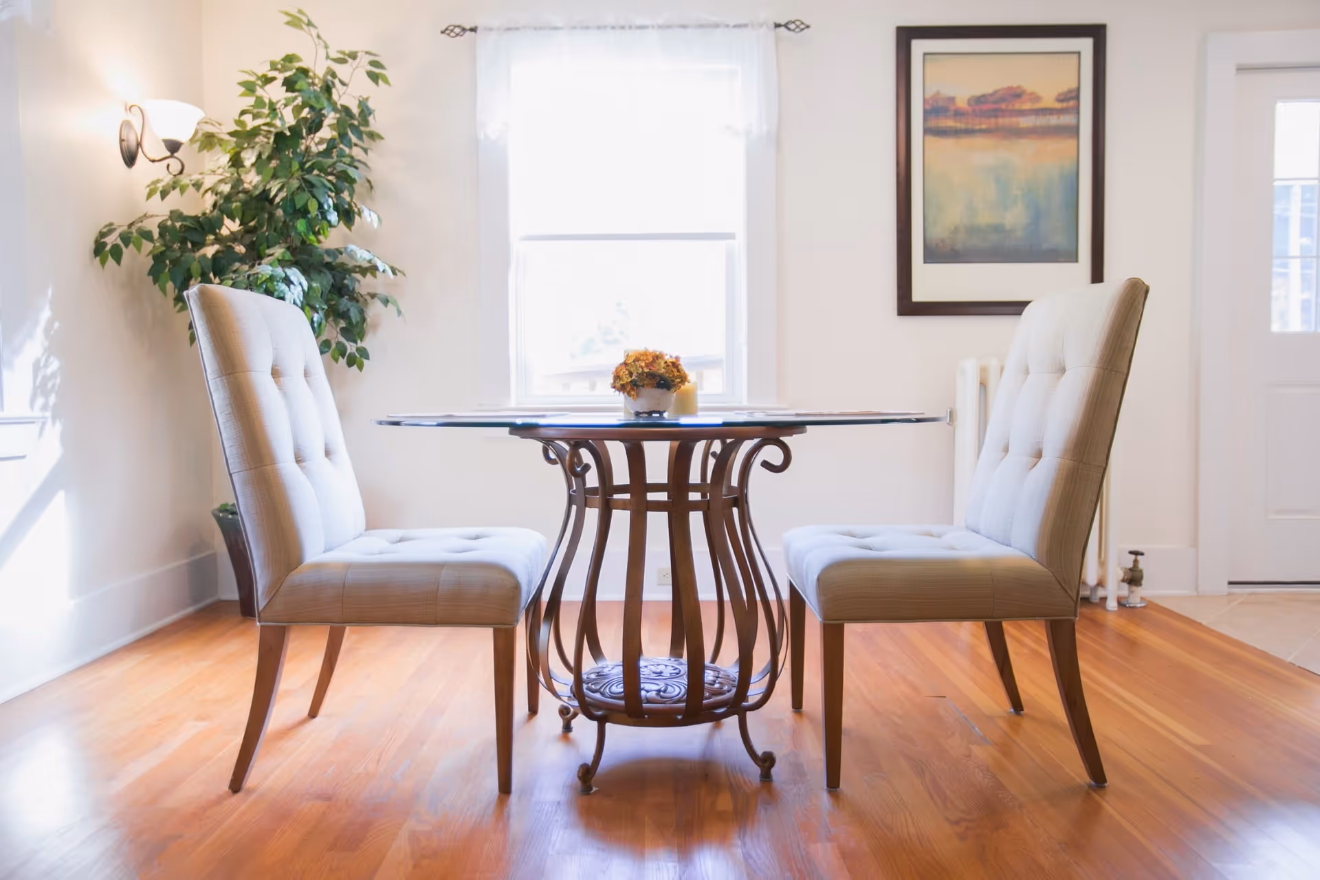 A bright dining area with a round glass-top table featuring an ornate metal base, flanked by two beige upholstered chairs. A potted plant is in the corner near a wall sconce, and a framed painting hangs on the wall next to a window with sheer curtains. The room has wooden flooring and white walls.
