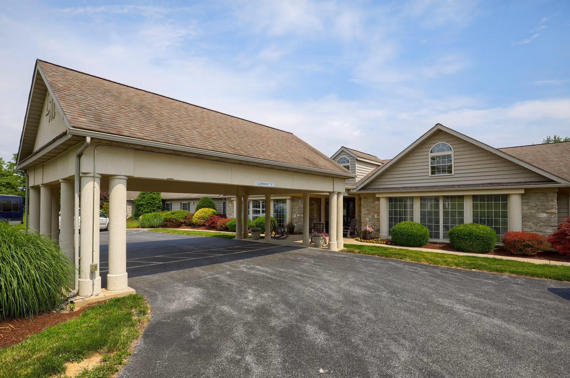Exterior view of a senior living facility with a covered driveway supported by columns, a paved entrance, and well-maintained landscaping including bushes and grass under a partly cloudy sky.