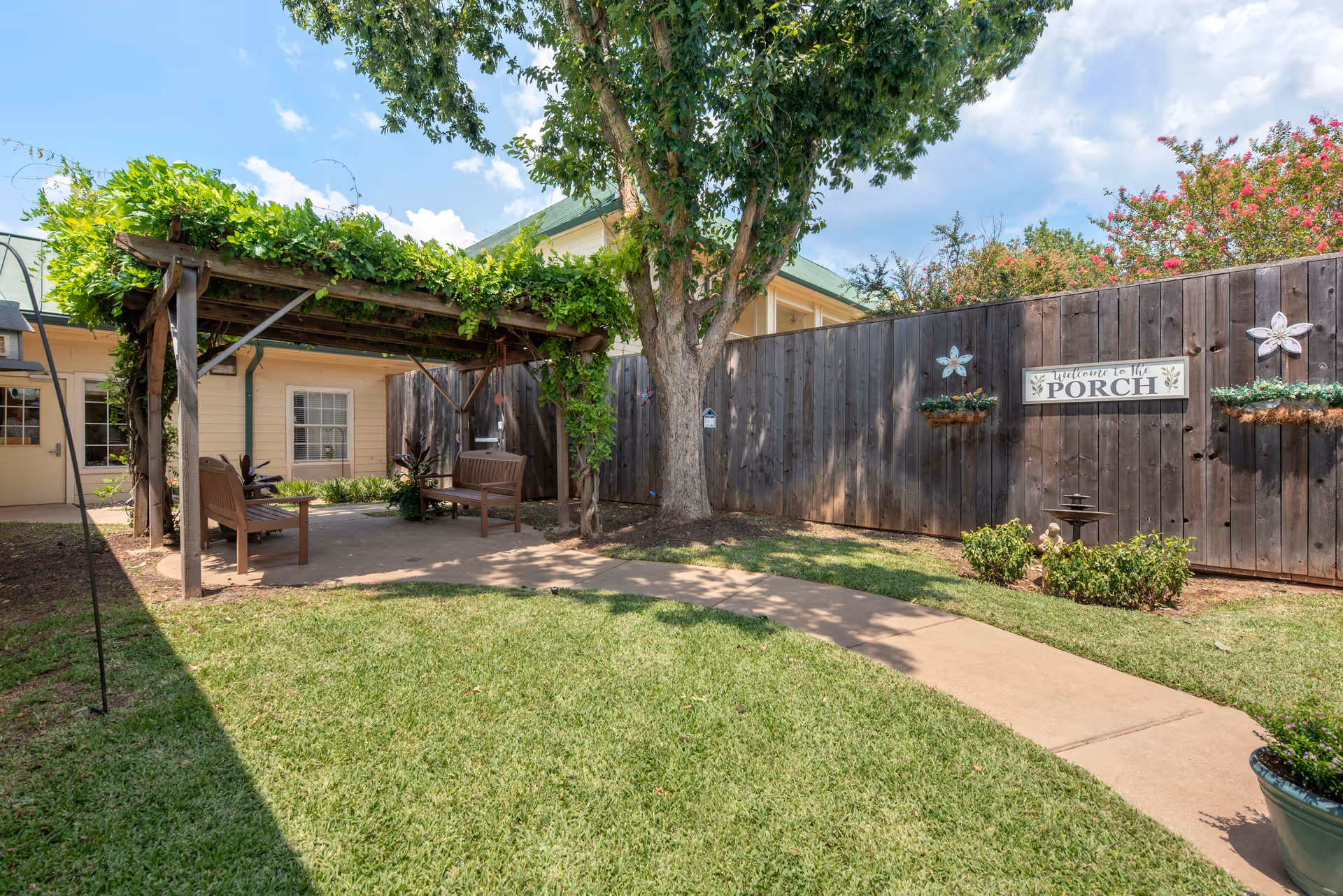 Outdoor garden area with a wooden pergola covered in green vines, two wooden benches underneath, a large tree, a wooden fence with a sign that reads 'Welcome to the Porch', and a concrete pathway surrounded by grass and small plants.