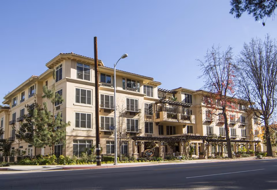 Exterior view of a multi-story senior living facility building with beige walls, multiple windows, balconies, and a pergola-covered entrance. Trees and greenery surround the building under a clear blue sky.