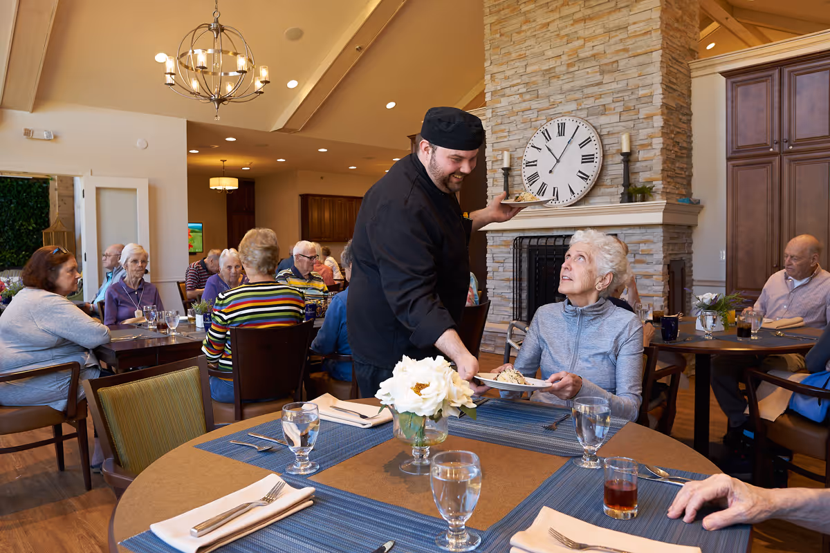 A dining room in a senior living facility where elderly residents are seated at tables with blue placemats, drinking glasses, and white napkins. A chef dressed in black is serving a plate of food to an elderly woman with short white hair sitting near a stone fireplace with a large clock above it. Other seniors are engaged in conversation or eating in the background.