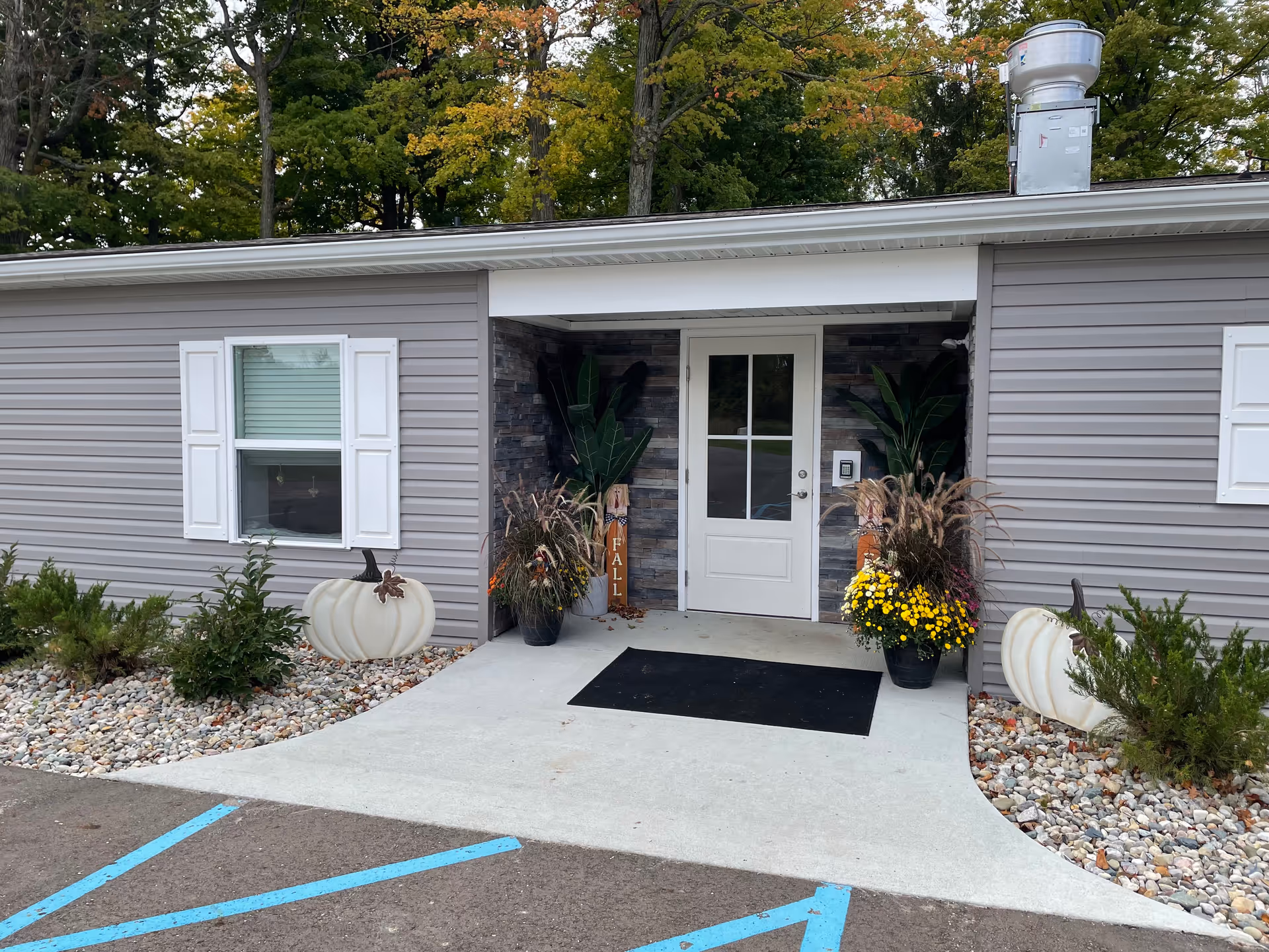 Entrance to a building with gray siding and white trim, featuring a white door with glass panels. The entrance is decorated with potted plants, yellow flowers, ornamental grasses, and white pumpkin decorations. There is a black doormat on the concrete walkway leading to the door, and a small sign with the word 'FALL' is visible among the plants. Trees with green and orange leaves are in the background.