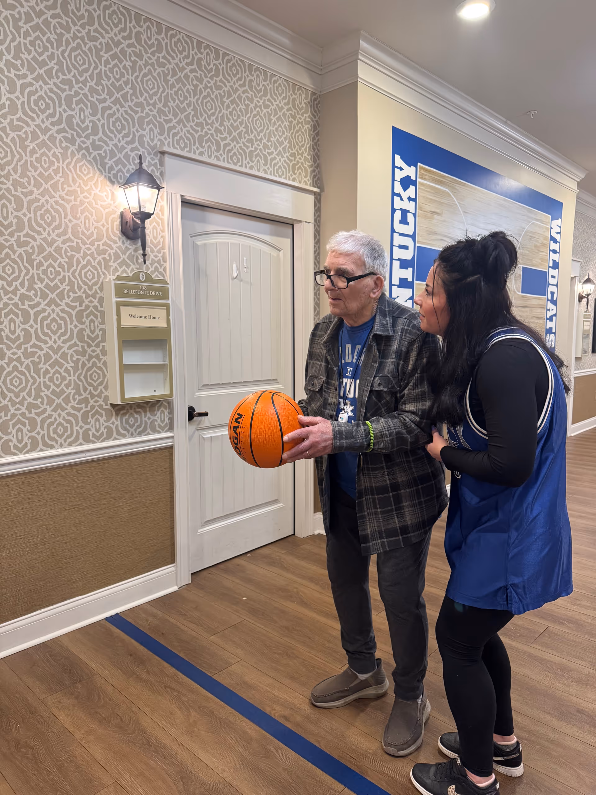 An elderly man holding a basketball is standing next to a woman in a blue sports jersey inside a hallway decorated with patterned wallpaper and a large Kentucky Wildcats basketball mural. The man is wearing glasses and a plaid shirt, and the woman is leaning slightly towards him, appearing to encourage or assist him.