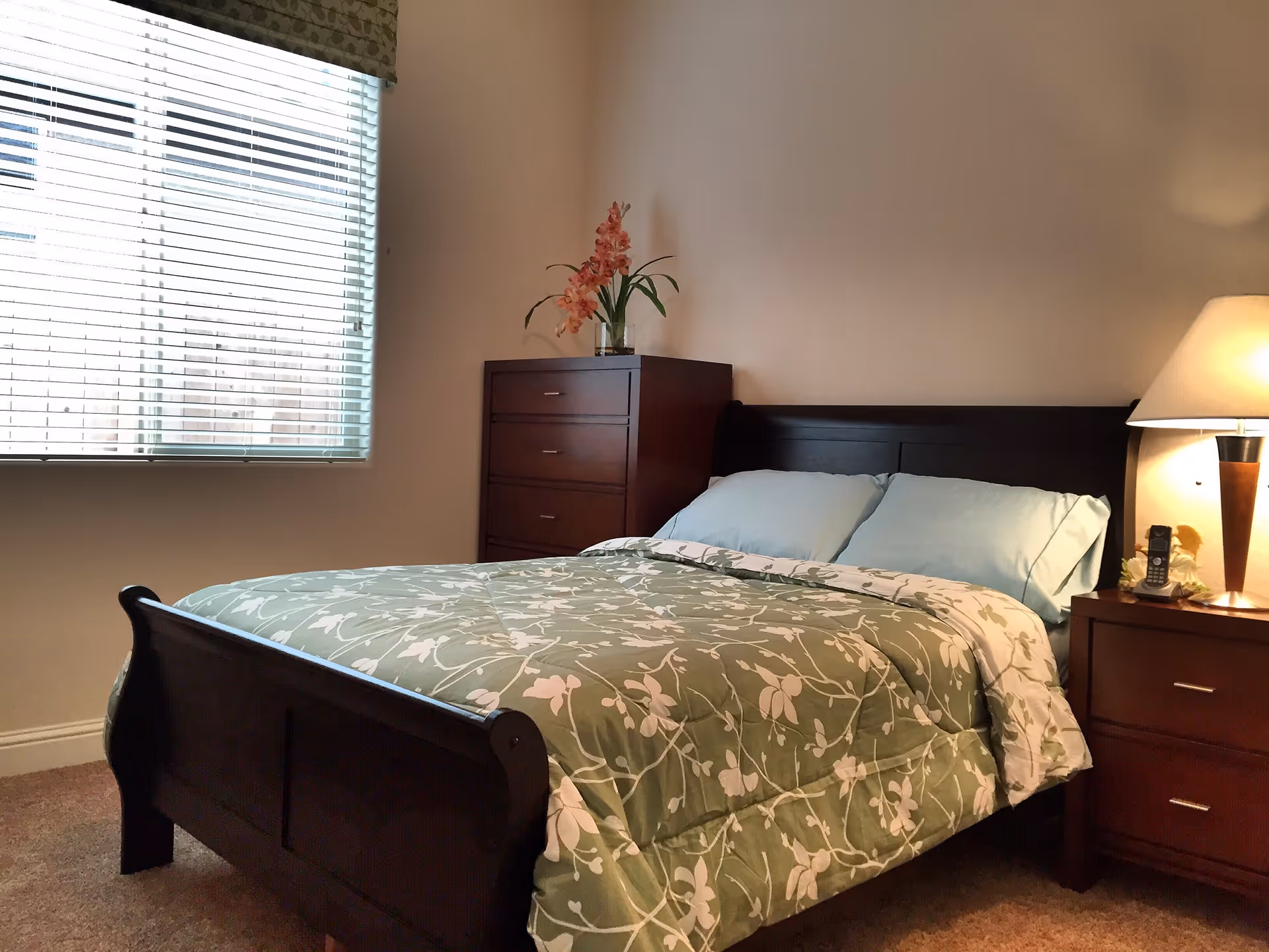 A bedroom with a dark wooden bed frame, a green floral bedspread, and light blue pillows. There is a wooden dresser with a vase of pink flowers on top, a nightstand with a lamp and a cordless phone, and a window with white blinds letting in natural light.