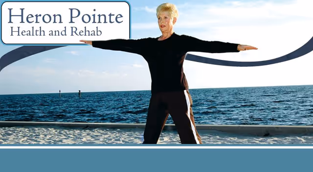 An elderly woman standing on a sandy beach near the ocean with her arms extended horizontally, wearing a black long-sleeve top and dark pants. The sky is clear with a few clouds, and there is a Heron Pointe Health and Rehab logo in the top left corner.