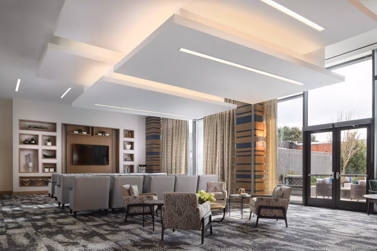 A modern senior living facility lounge area with multiple gray armchairs arranged in rows facing a wall-mounted flat screen TV. The room features large floor-to-ceiling windows with beige curtains, patterned carpet, and unique ceiling light panels. There are also a few patterned armchairs around small tables with decorative items and flowers. Outside the windows, an outdoor seating area is visible.