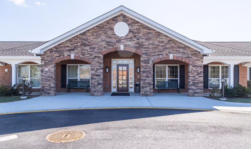 Front exterior view of a senior living facility with a stone facade, a central glass door entrance, two benches under covered archways on either side of the door, and a paved driveway in front.