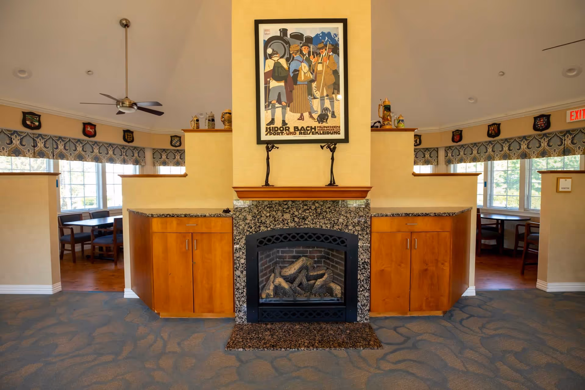 Interior view of a common area featuring a central fireplace with a granite surround and wooden cabinets on either side. Above the fireplace is a framed vintage poster depicting people in traditional attire. The room has carpeted flooring, ceiling fans, and windows with patterned valances. Two dining areas with tables and chairs are visible through openings on both sides of the fireplace.