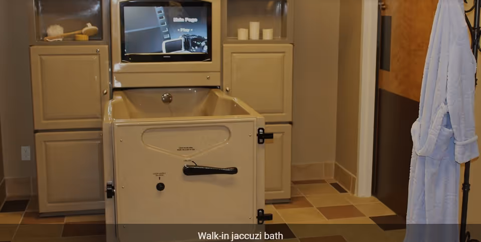 A walk-in jacuzzi bath with a control panel and a screen above it, surrounded by beige cabinets and tiled flooring. A white bathrobe hangs on a stand to the right.