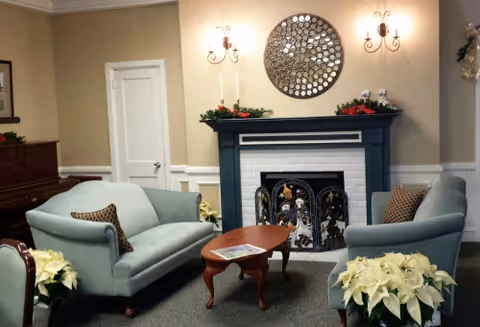 A cozy living room area with two light blue upholstered sofas facing each other across a wooden coffee table. The room features a white brick fireplace with a decorative black screen and holiday decorations on the mantel. Above the fireplace is a round mirror with a circular pattern. There are wall sconces on either side of the mirror, and white poinsettia plants are placed near the sofas.