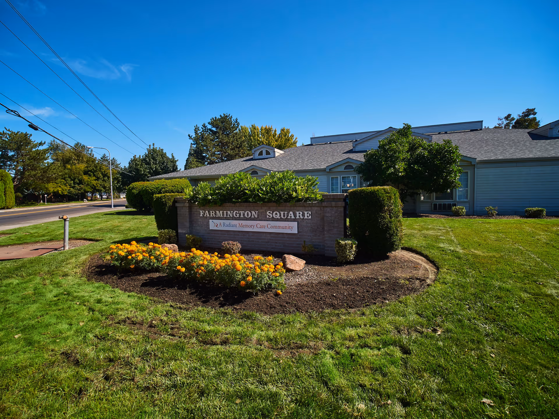 Exterior view of Farmington Square Medford facility with a landscaped lawn, flower bed with orange flowers, trimmed bushes, and a sign that reads 'Farmington Square A Radiant Memory Care Community' under a clear blue sky.