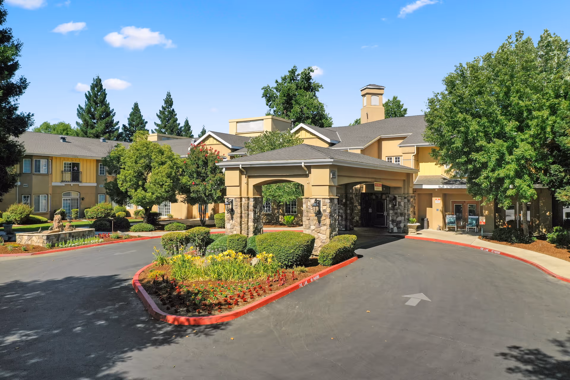 Exterior view of Quail Park on Cypress senior living facility showing the main entrance with a covered driveway, surrounded by landscaped greenery and trees under a clear blue sky.