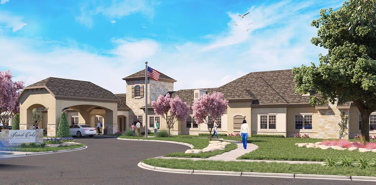 Front exterior view of French Oaks Memory Care facility with a driveway, landscaped lawn, pink flowering trees, an American flag on a flagpole, and several people walking near the entrance under a covered drop-off area.