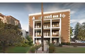 Front exterior view of a three-story brick building with balconies on each floor, surrounded by trees and greenery under a partly cloudy sky.