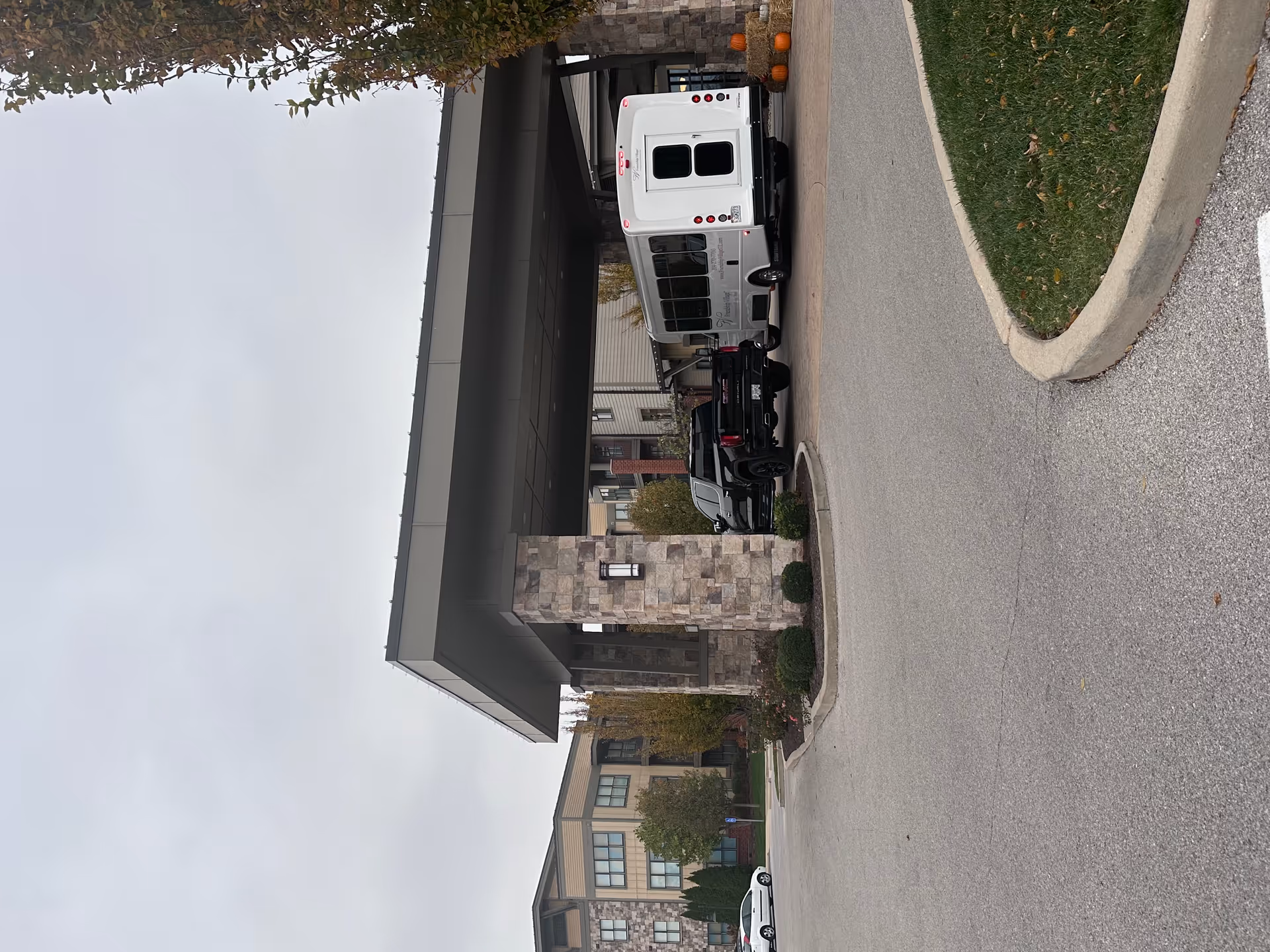 Entrance area of Friendship Village Sunset Hills featuring a covered drop-off zone with stone pillars, a black pickup truck, and a white shuttle bus parked underneath. The background shows a multi-story residential building with large windows and autumn trees.