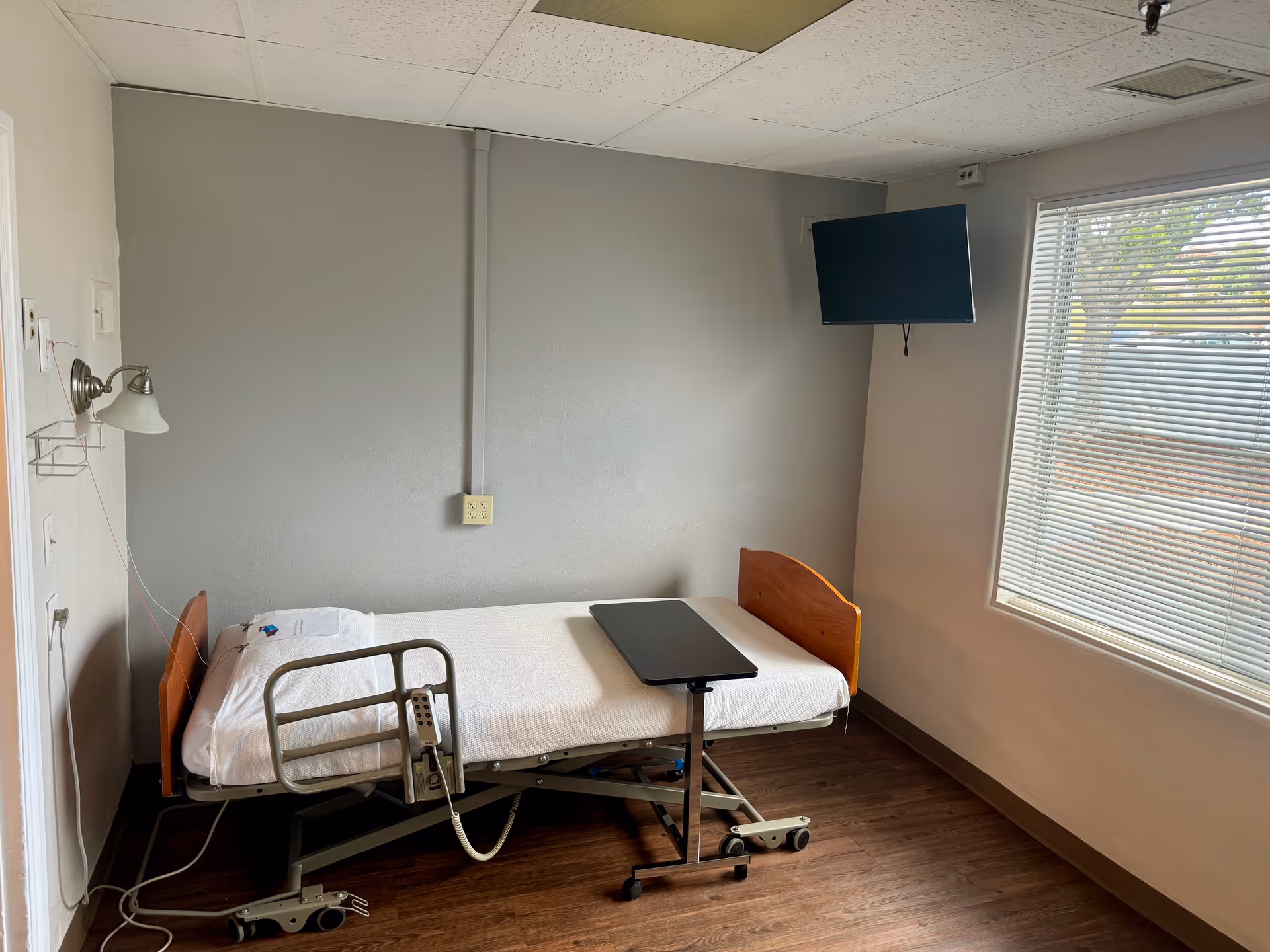 A small, simple room with a hospital-style bed featuring a white blanket and a wooden headboard and footboard. There is a movable overbed table on wheels positioned over the bed. A wall-mounted lamp is on the left wall above the bed, and a flat-screen TV is mounted on the wall near a window with blinds, letting in natural light. The floor is wood, and the walls are painted light gray and white.
